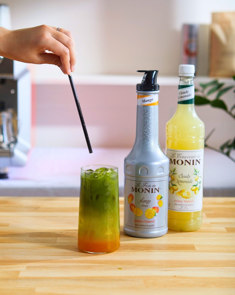 Layered mango matcha lemonade beside MONIN mango and cloudy lemonade bottles